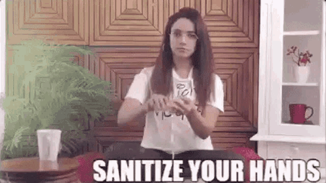 a woman is sitting at a table in a living room with a cup of coffee and a sign that says sanitize your hands .