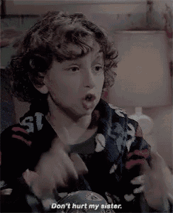 a young boy with curly hair is sitting on a bed and talking to his sister .