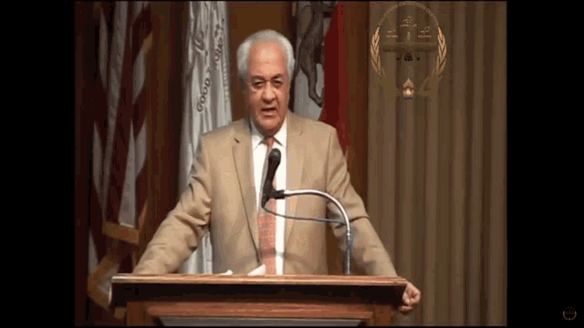 a man in a suit and tie stands at a podium speaking into a microphone in front of flags