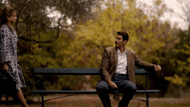 a man in a suit sits on a park bench while a woman walks by
