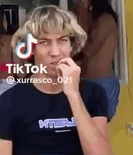 a man with blonde hair is making a funny face with his finger in his mouth and a tiktok logo above his head .