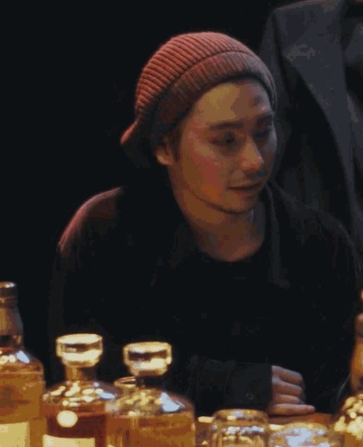 a man wearing a red beanie sits at a table with bottles of alcohol