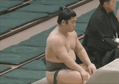 a sumo wrestler is sitting on a mat in a stadium while another man sits behind him .