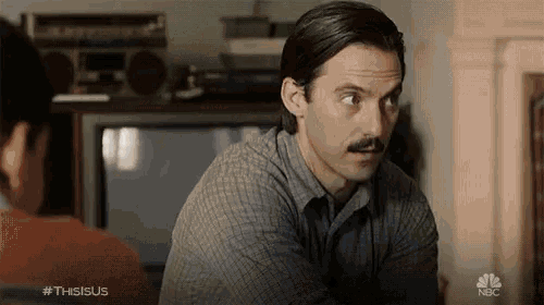 a man with a mustache is sitting in front of a television with the nbc logo on the bottom