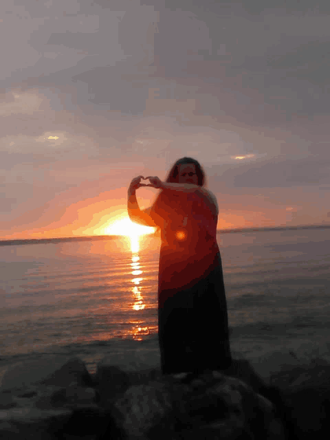 a woman making a heart shape with her hands in front of a sunset over the ocean