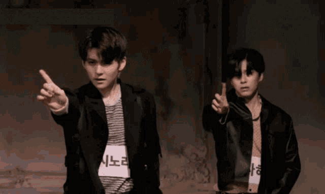 two young men are standing next to each other in a dark room and one is pointing at the camera .