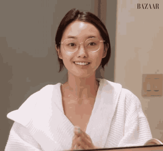 a woman wearing glasses and a white robe looks at herself in the mirror .
