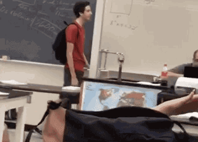 a man with a backpack is standing in front of a chalkboard in a classroom .