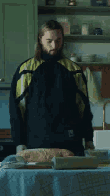 a man with long hair and a beard is standing in a kitchen with bread on a table .