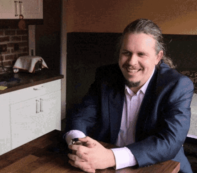 a man in a suit sits at a table with his hands folded and smiles