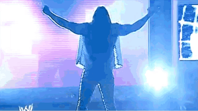 a man with long hair is standing on a stage with his arms outstretched in front of a screen that says wrestling .