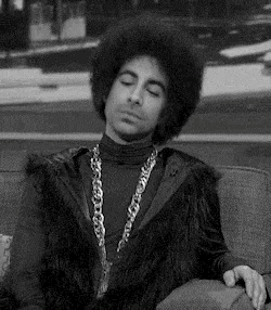 a black and white photo of a man sitting on a couch wearing a fur vest and a chain .