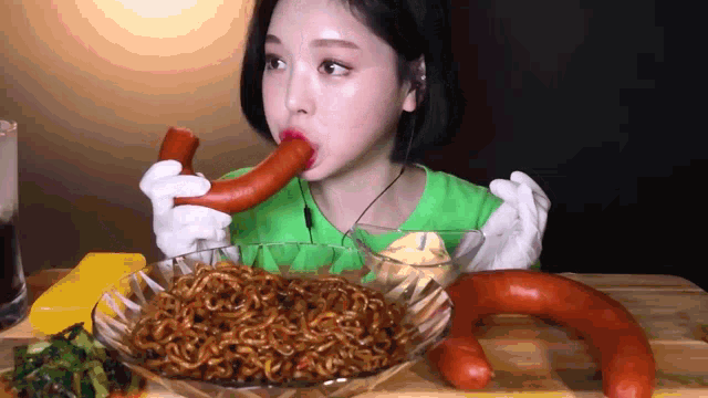 a woman in a green shirt is eating noodles with a sausage in her mouth