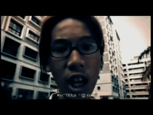 a man wearing glasses is standing in front of a building and says " xxx "