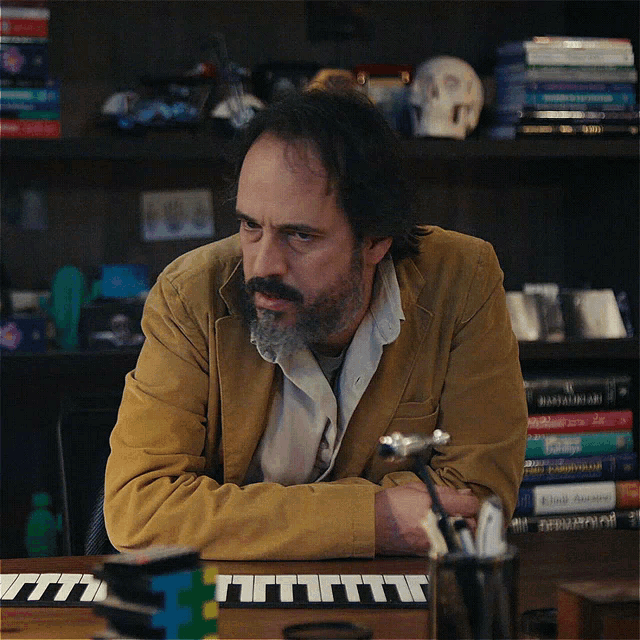 a man with a beard sits at a desk with a keyboard in front of him and a stack of books on a shelf