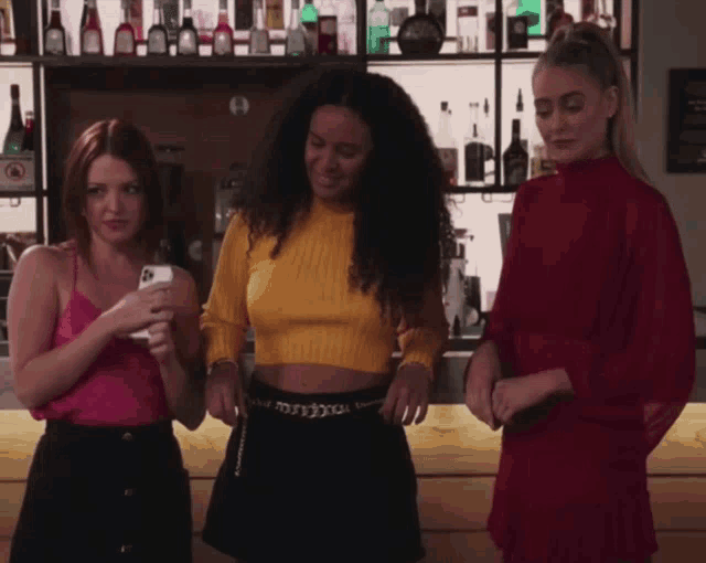 three women are standing in front of a bar with bottles on the shelf behind them