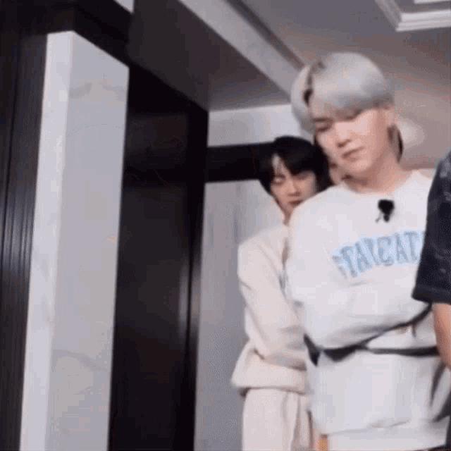 two men are standing next to each other in a hallway . one of the men is wearing a straitjacket .