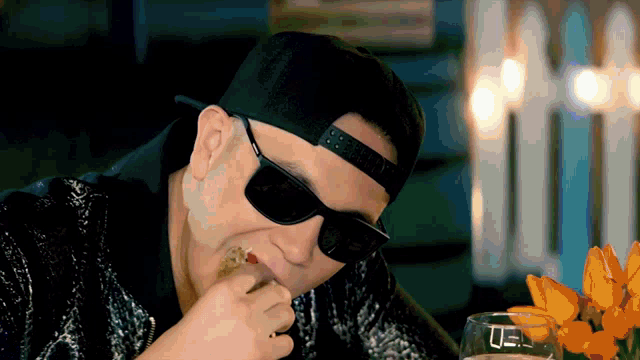 a man wearing sunglasses and a baseball cap is eating a sandwich