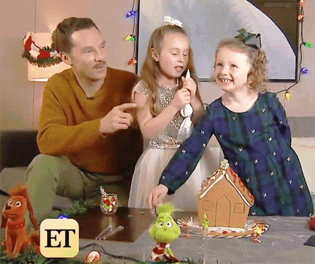 a man and two little girls are sitting at a table with a gingerbread house in front of them and an et logo