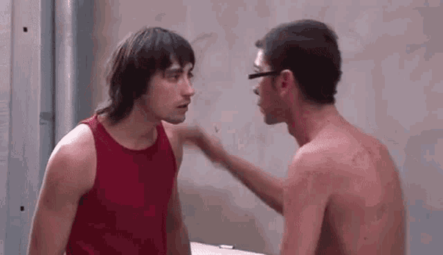two shirtless men are standing next to each other and talking to each other . one of the men is wearing glasses .
