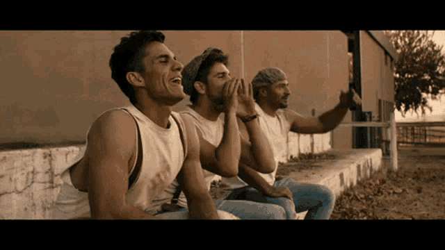three men are sitting on a ledge laughing and one has a hat on