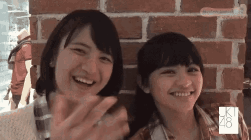 two girls are smiling in front of a brick wall with the letters jk48 on it