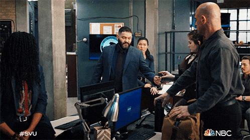 a group of people standing around a desk with a nbc logo on the bottom right