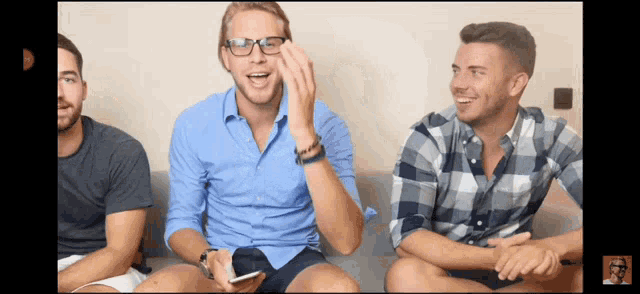 three men are sitting on a couch laughing and one has glasses on