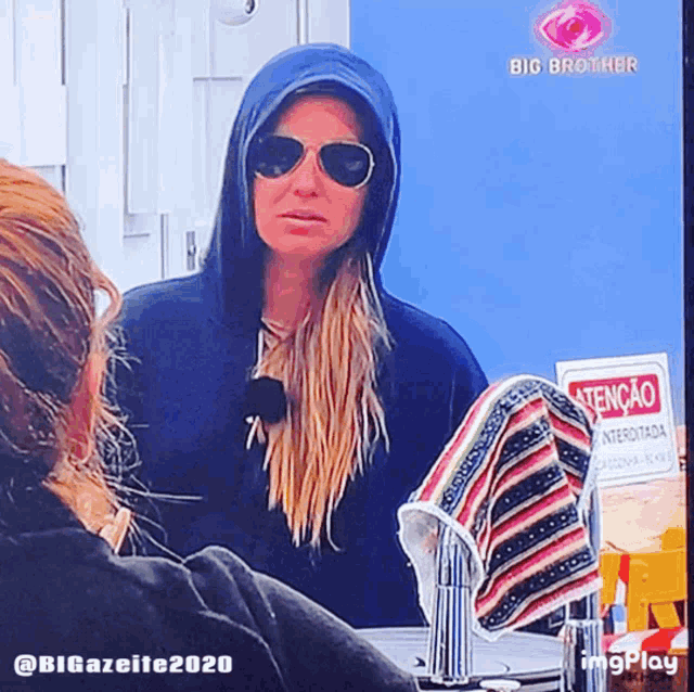 a woman wearing a hoodie and sunglasses looks at herself in a mirror with big brother written on the screen behind her