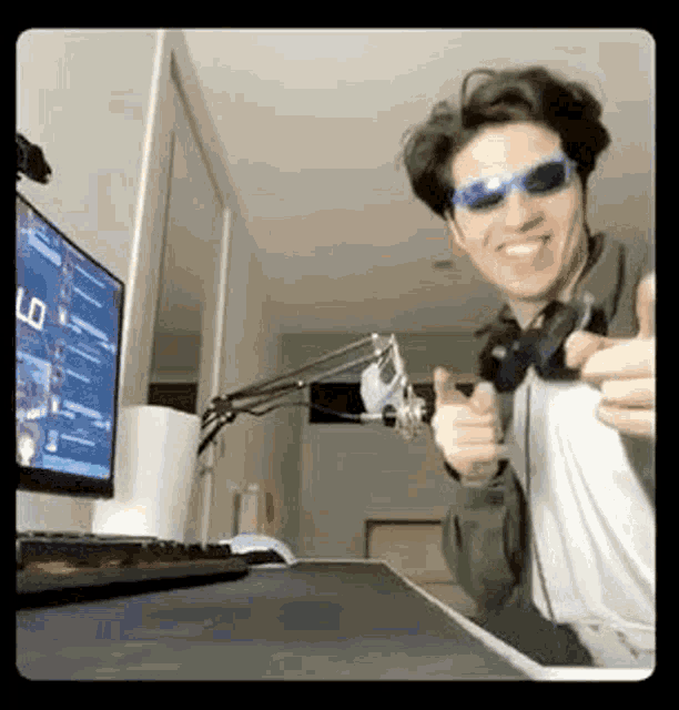a man wearing sunglasses and headphones is giving a thumbs up while sitting in front of a computer .