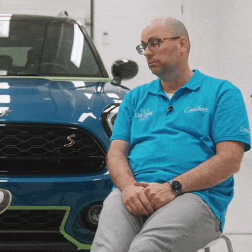 a man wearing a blue car care shirt sits in front of a car