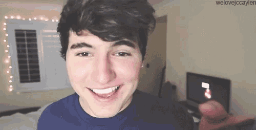 a young man in a blue shirt is smiling and pointing at the camera in a bedroom .