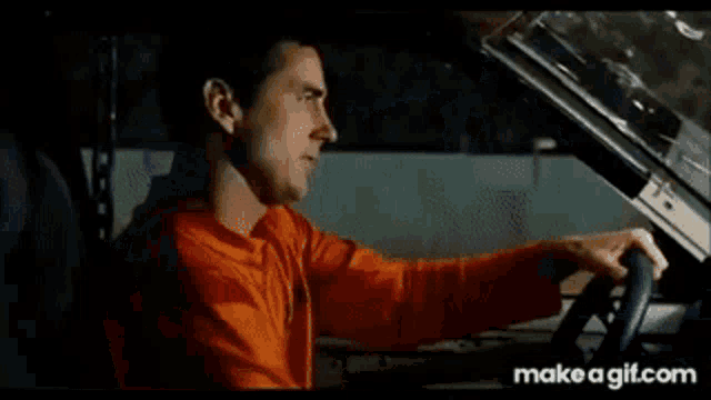 a man in an orange shirt is driving a car with his hands on the steering wheel .