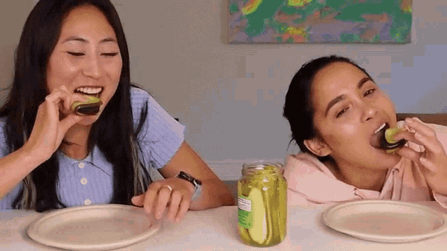 two women are sitting at a table eating pickles with a jar of pickles next to them .