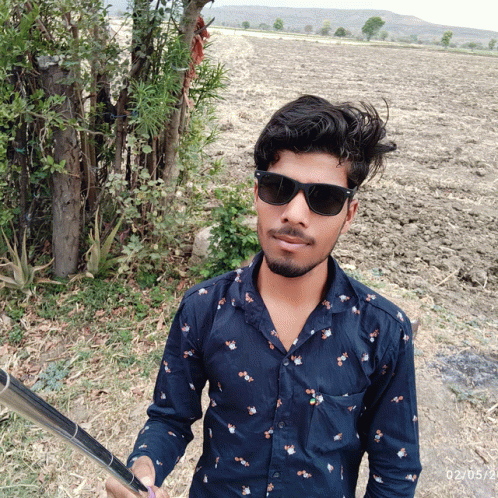 a young man wearing sunglasses and a blue shirt stands in a field