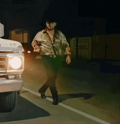 a man in a cowboy hat is walking down a street next to a truck at night .