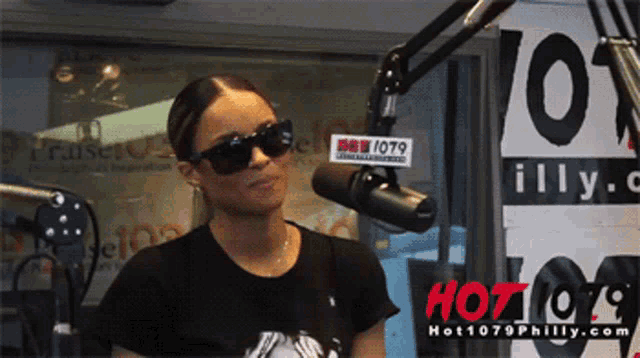 a woman wearing sunglasses stands in front of a microphone in front of a sign that says hot1079philly.com