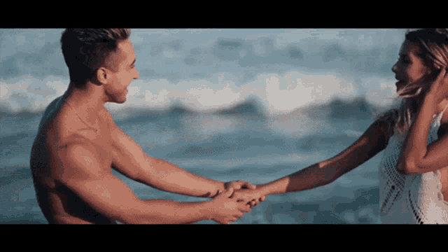 a shirtless man and a woman holding hands on a beach