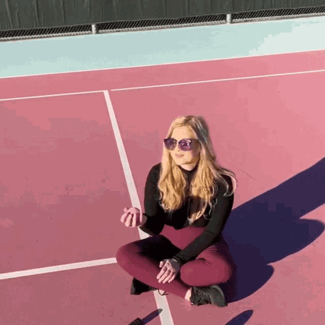 a woman wearing sunglasses sits on the ground on a pink tennis court