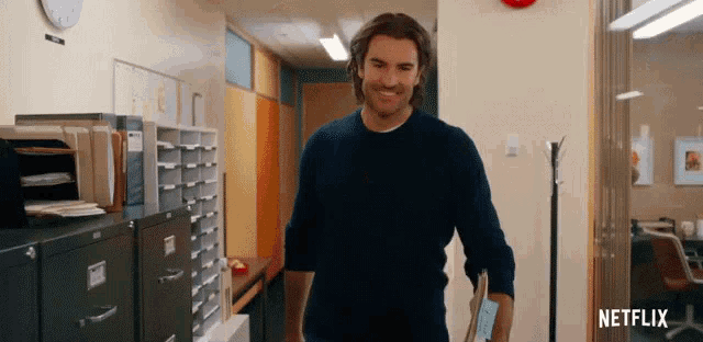 a man in a blue sweater is walking in a hallway with netflix written on the wall
