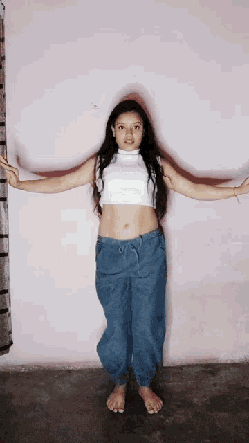 a girl in a white crop top and blue pants is standing in front of a pink wall