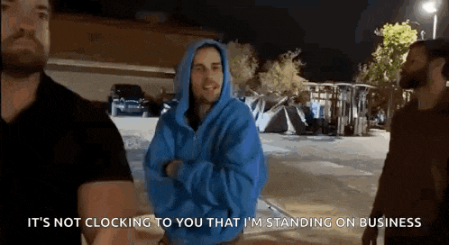 a man in a blue hoodie is standing in a parking lot talking to another man .