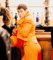 a man in an orange suit is standing in front of a bar holding a red bag .