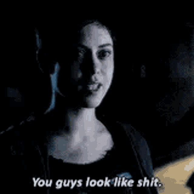 a woman is talking to a man in a dark room and saying `` you guys look like shit . ''