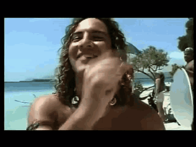 a shirtless man with long hair is standing on a beach with a surfboard .