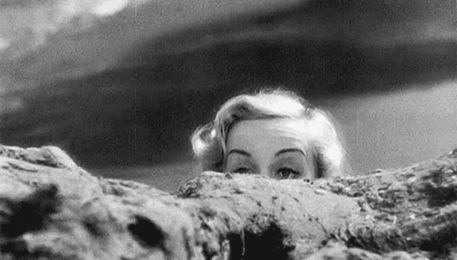 a black and white photo of a woman peeking over a rock .