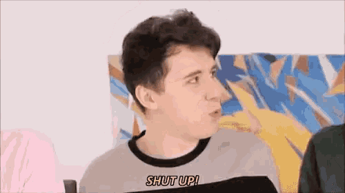a young man is sitting in front of a painting and talking to someone while wearing a shut up shirt .