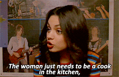 a woman is talking in front of a poster that says the woman just needs to be a cook in the kitchen