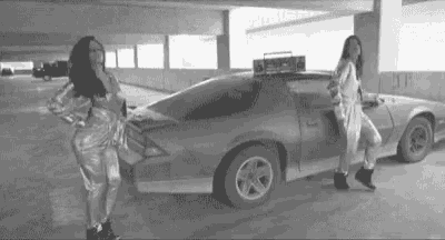 two women are dancing in front of a car in a parking garage .