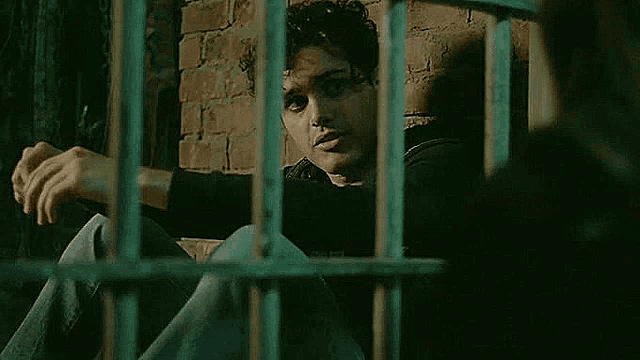 a man is sitting in a jail cell behind bars looking out .